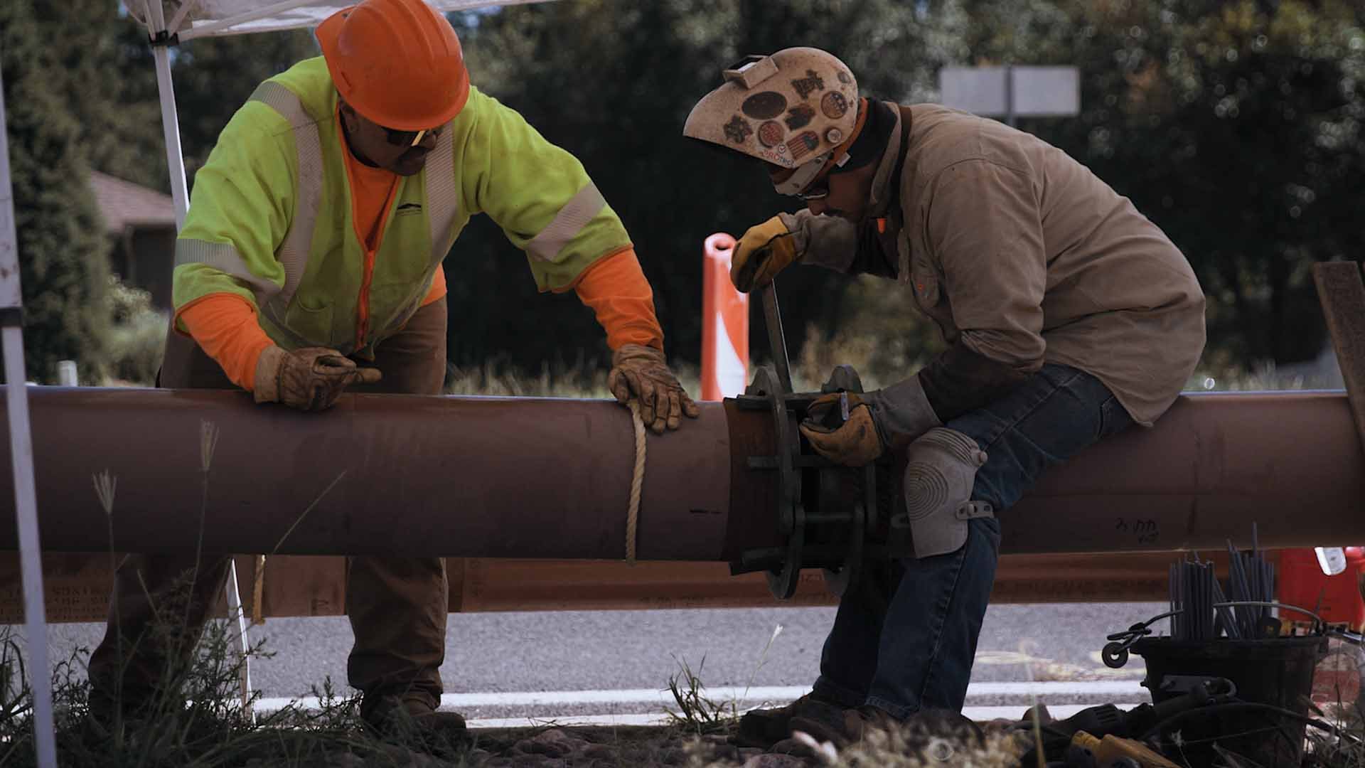 Two wastewater construction workers stand and sit near a large iron pipe