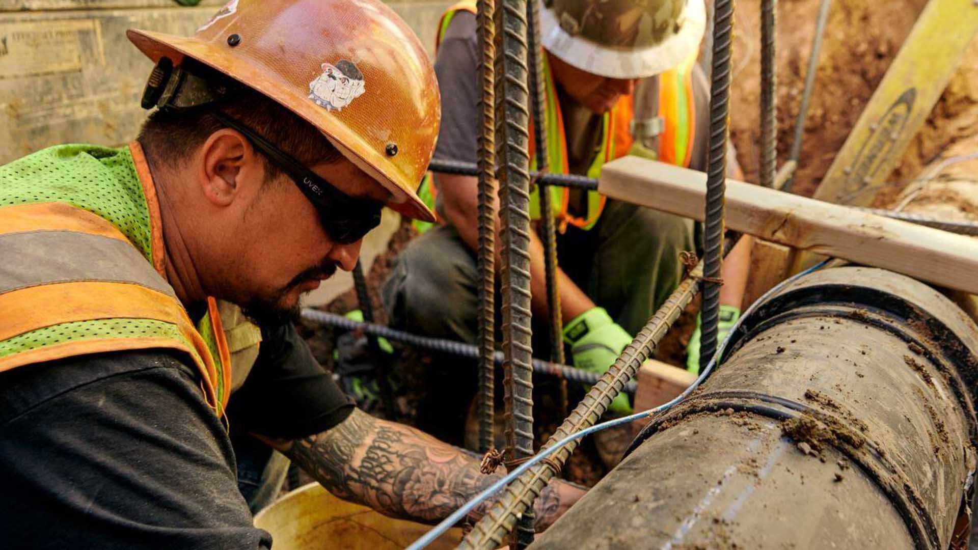 Two construction workers wearing hard hats handle rebar around a steel pipe.