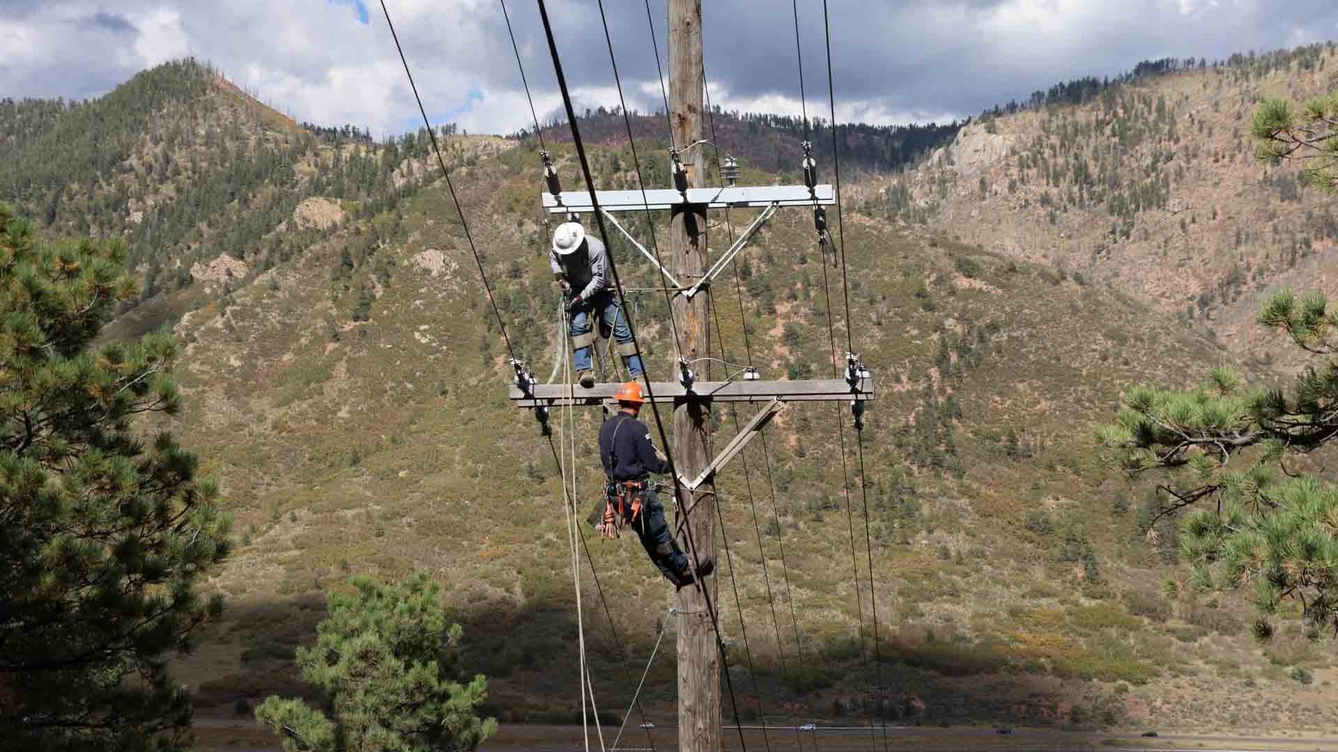 Two line workers are perched high on a power pole near lines with mountains in the background.