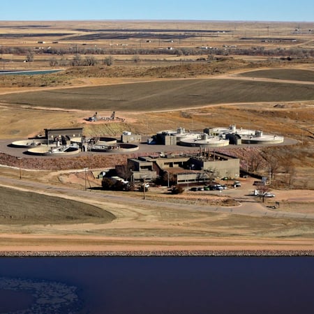 An aerial photo of a tan brick facility with circular structures emerging from a hill behind the main building. In the foreground, a dark blue lake is visible before a road. It's a sunny day and the surrounding fields are brown and tan, indicating this photo was taken in winter.