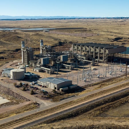 Aerial image of Front Range Power Plant. It features two smokestacks, which are not very prominent. There are overhead lines and substation equipment next to the plant and a vast array of industrial equipment next to the stacks. The surrounding landscape is brown grass.