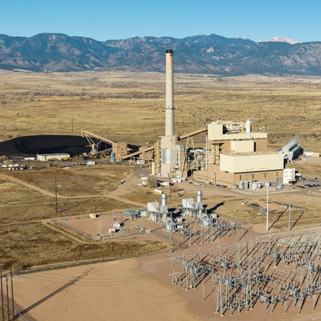 An aerial video of Nixon Power Plant. One large smokestack is visible to the left of a large building. A coal pile is visible behind the plant and a substation is visible in the foreground. Mountains and the very summit of Pikes Peak are visible in the background.