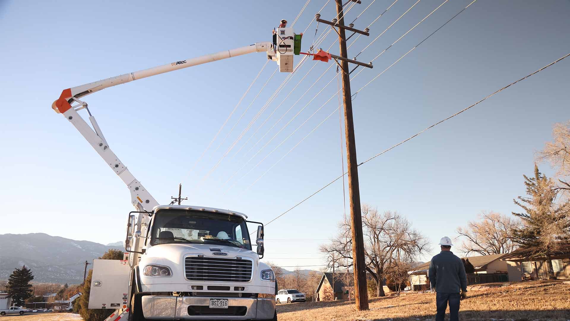 A line worker stands in a bucket extended from a truck next to a wooden power pole.