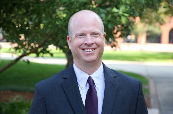 A photo of Tristan Gearhart. He is wearing a suit and smiling. There is a tree and a sidewalk in the background.