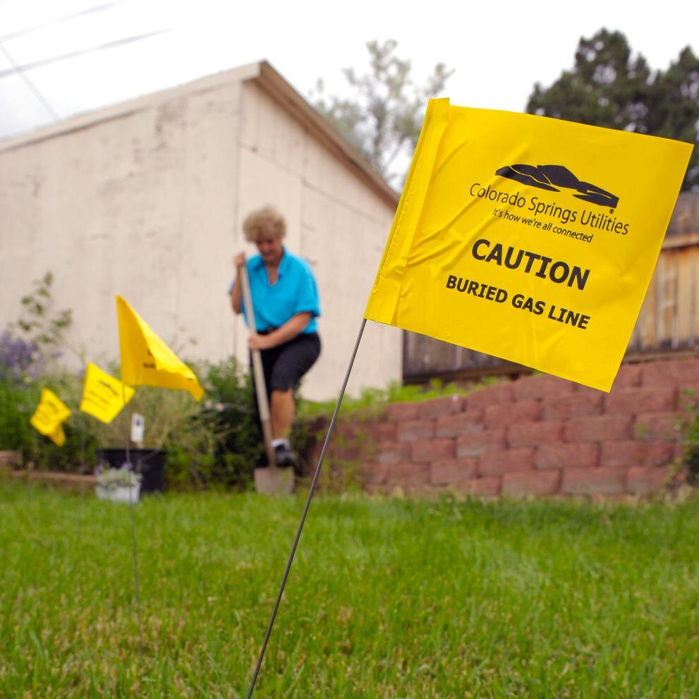 A back yard with small yellow flags in it, with the words "CAUTION BURIED GAS LINE" are printed on the flags. A woman in a blue shirt is digging. 