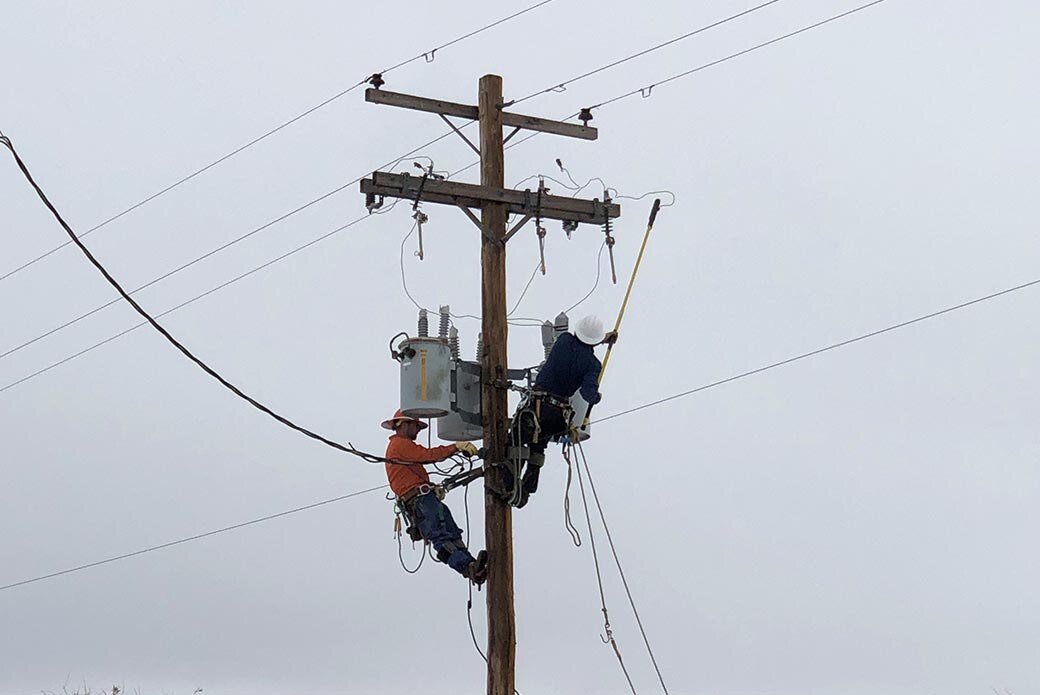 Two workers using climbing harnesses to scale a wooden utility pole. One of them is holding a yellow pole and the other is using a tool on a wire. 