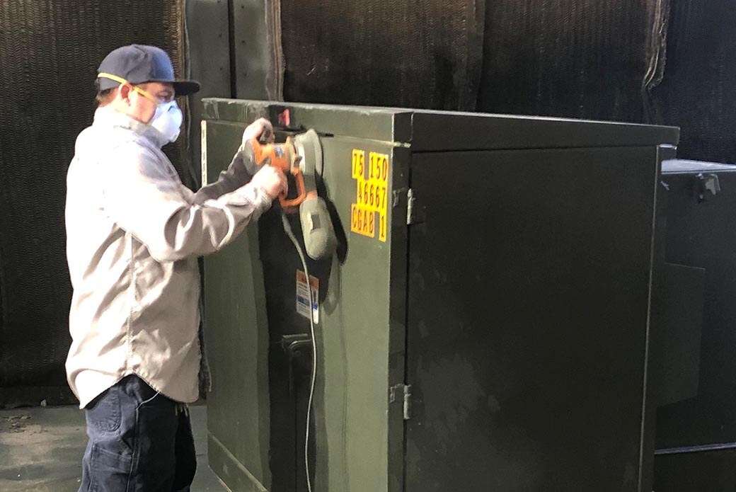 A man wearing a dust mask using an electric rotary tool on the panels of a large transformer.