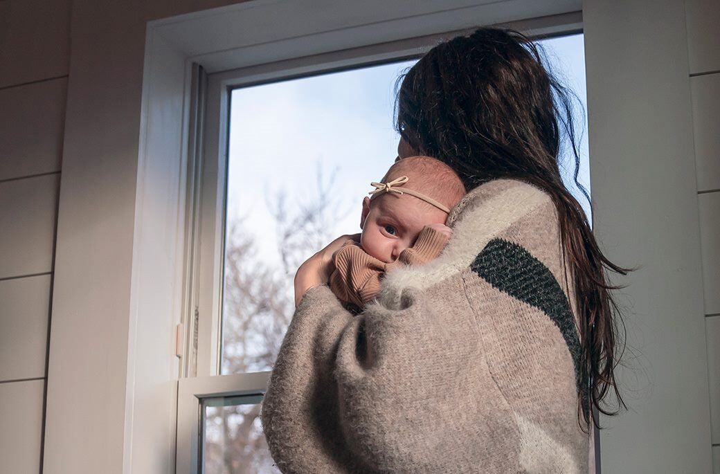 A woman wearing a cardigan and holding a baby close to her chest. She is looking out a window.  A tree appears outside the window.