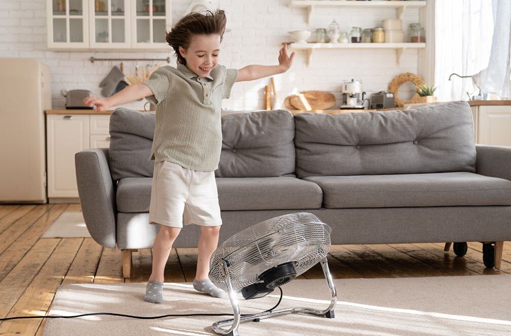 A small, smiling child wearing a short sleeve shirt, shorts, and socks in a living room with his arms stretched out while a fan blows air onto him. 