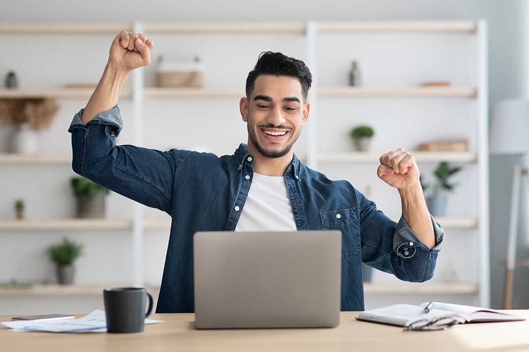 A man sitting at a laptop on a table with his fists in the air and smiling. A notebook, some papers, a pair of glasses, and a mug sit alongside him. 