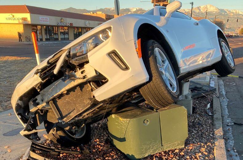 A white car crashed into a small residential transformer. The front of the car is smashed and car is leaning to the right on top of the transformer