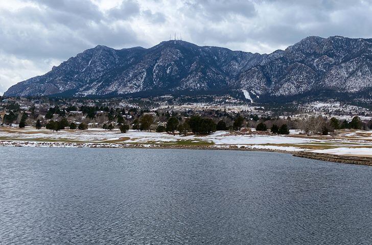 A landscape image of a lake with grass covered ground and snow surrounding it. The Cheyenne Mountains are in the background. The sky is cloudy. 