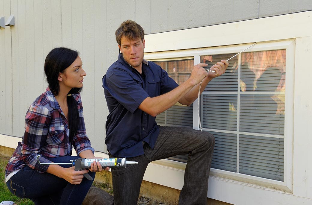 Person removes old caulking from window while another kneels nearby holding a caulk gun to apply a new bead.