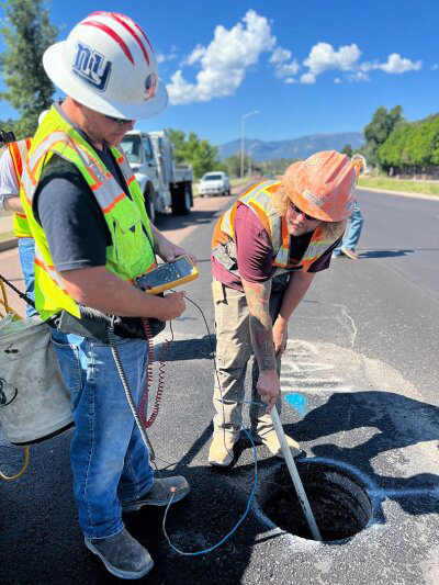 Two individuals in safety gear stand looking at a hole in a street. One of the workers lowers a long pvc pole into the hole. Mountains are visible in the distance, it's a sunny day.