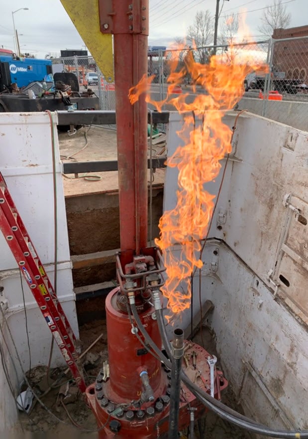 A piece of red gas line equipment is exposed out of the ground. Flames are erupting from the end of of one of the gas lines.