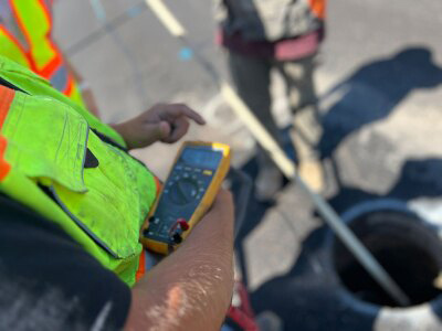 Person in safety vest stands near a hole in a street, which is slightly out of focus. The person is holding an instrument with a display, it appears to be a voltmeter. The focus is on the person's hand.