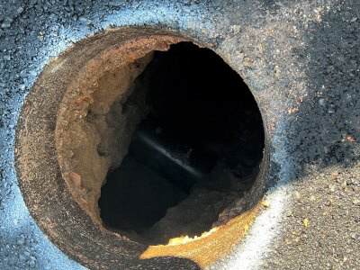 A circular hole in pavement. The hole is approximately 15 feet deep, and a white pipe is barely visible at the bottom of the hole. White spray paint surrounds the outer rim of the hole. 