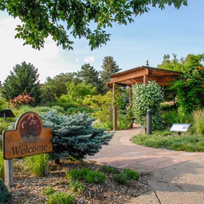 A welcome sign at the entrance at the Conservation and Environmental Center. A paver pathway leads toward a wooden pergola surrounded by lush greenery