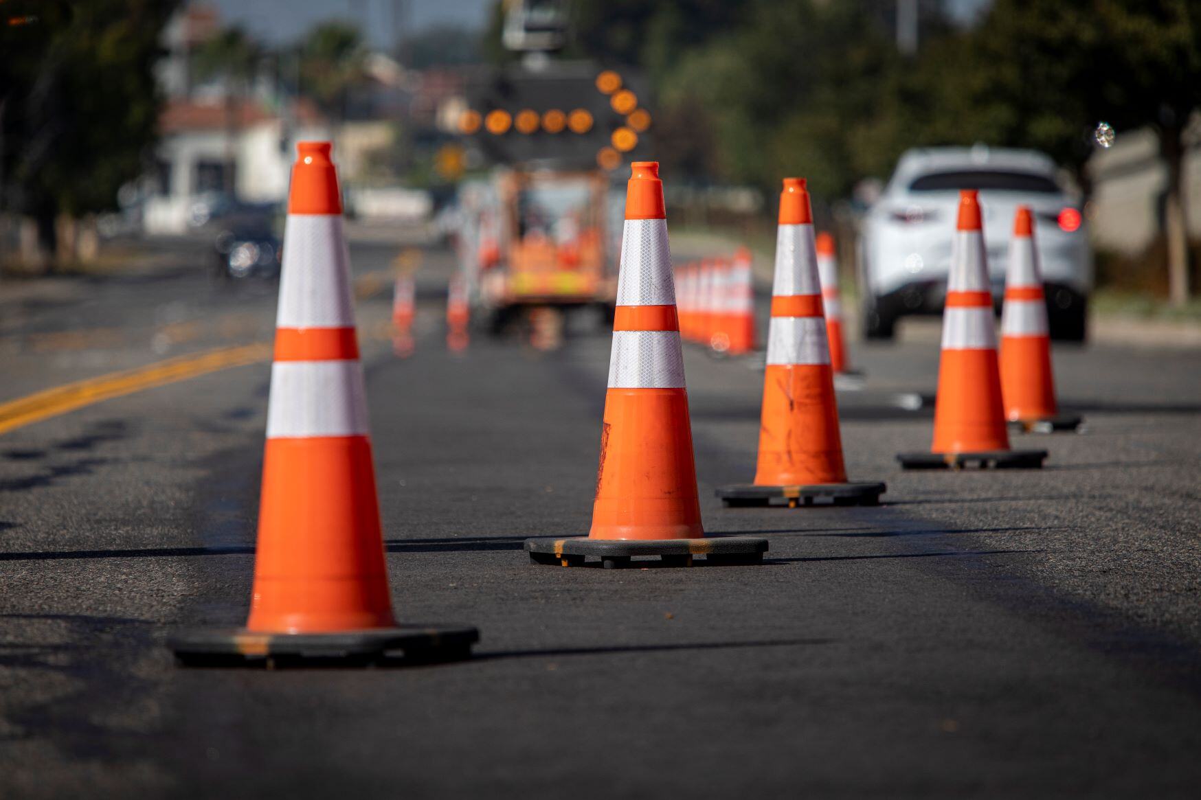 A series of orange traffic cones  placed on a street, leading toward construction vehicles with warning lights, indicating a work zone ahead.