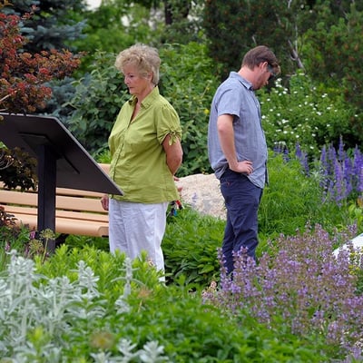 Two individuals stand in a garden with purple & green plants and flowers. One is looking at a display board, the other looks at the flowers.