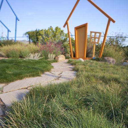 A stone pathway leads through a grassy area to an oversized, open orange frame in the shape of a house without walls or roof next to purple flowers.