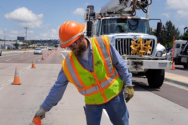 Man in orange hard hat and neon safety vest putting out traffic cones in busy street with cars and utility truck behind him. 