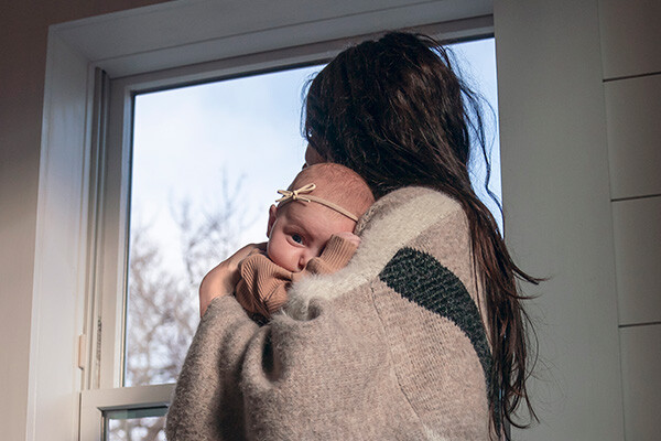 A small baby is held by a young woman who is wearing a warm sweater as she looks out the window