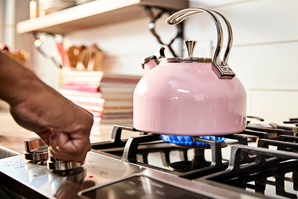 A hand turning on natural gas with a pink tea kettle on the burner and blue natural gas flame underneath