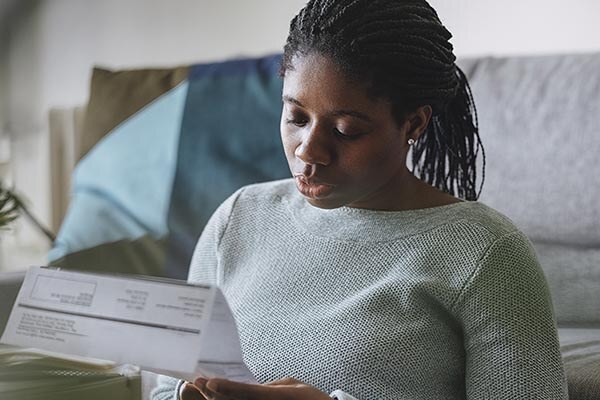 A young woman in green shirt sits on her couch and looks at a bill with a neutral expression on her face