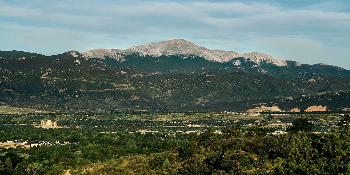 A scenic photograph of Colorado Springs with lush green trees, Garden of the Gods in the right side of the frame with mountains in the background. 