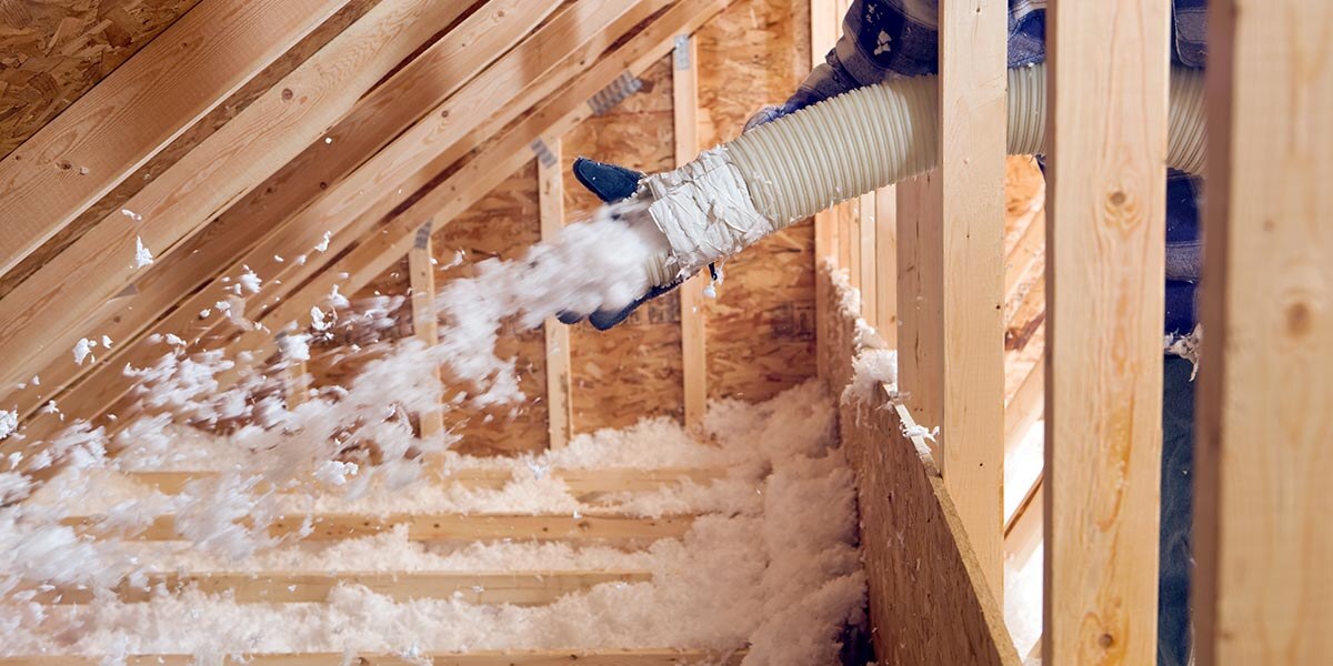 A person is shooting insulation into a new attic via a white hose. They are wearing a blue flannel shirt and blue gloves. 