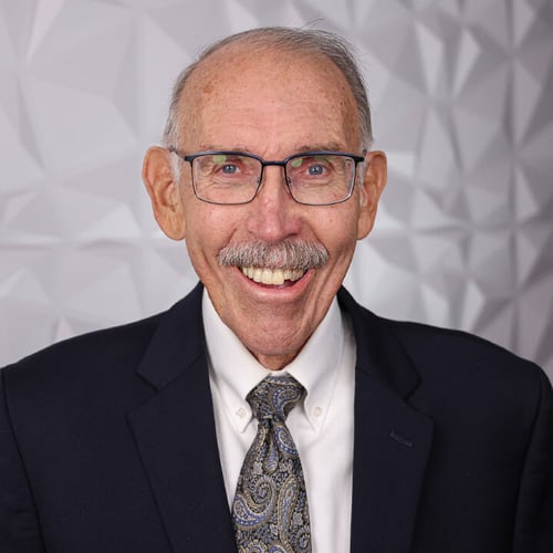 A headshot of an elderly man named David Watson. He is wearing a black suit, glasses and smiling in front of a white textured wall. 