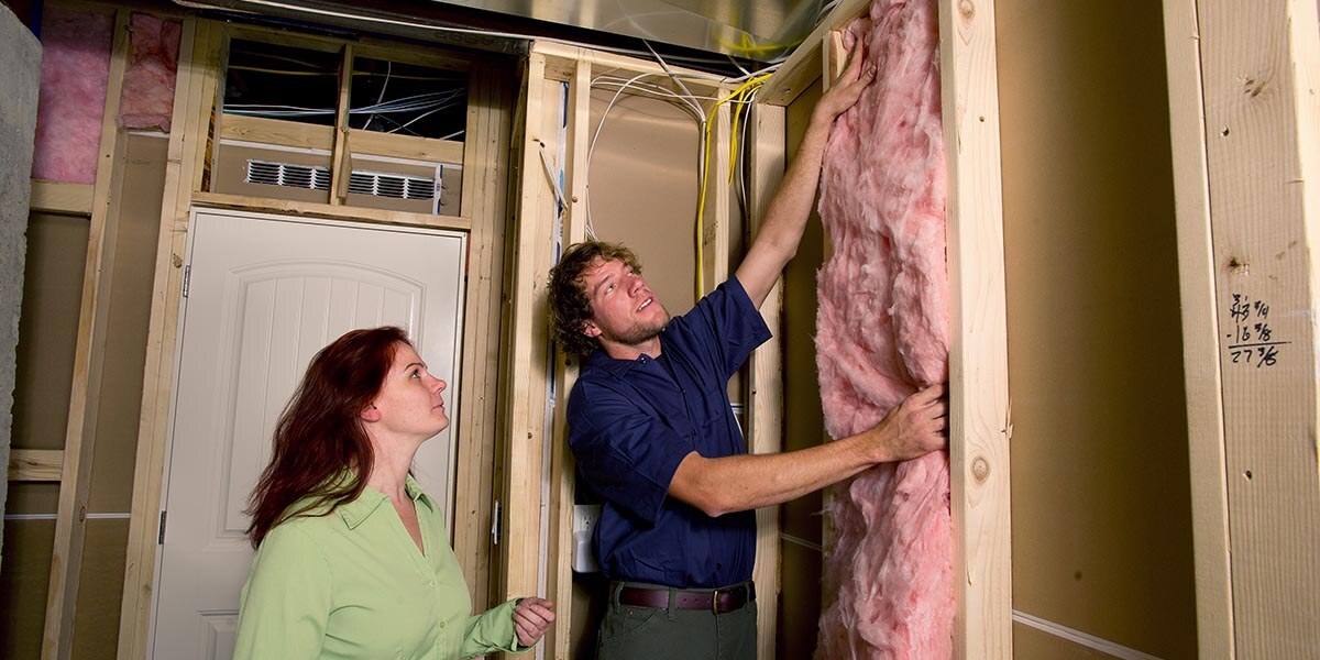 A man and woman installing pink insultation in an unfinished room. The man is wearing a blue shirt and the woman is wearing a light green shirt. 