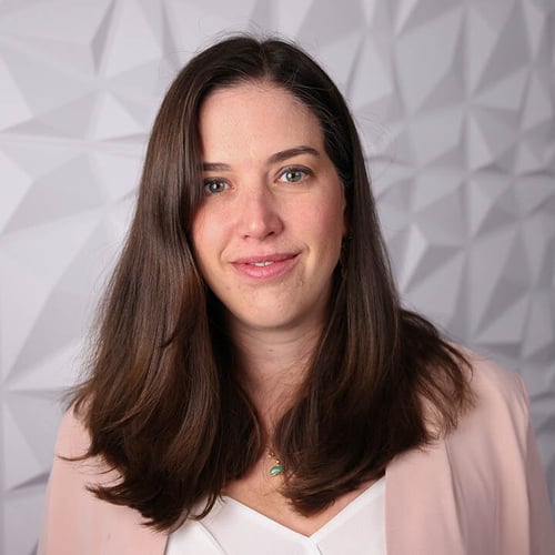 A photograph of a woman named "Kate Danner". She is standing in front of a white textured wall and is wearing a light pink shirt. 