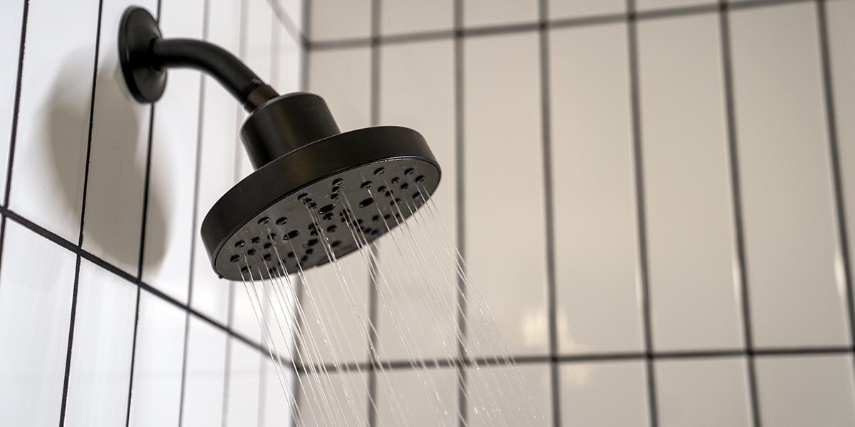 A photograph of a black showerhead being used in a shower. The walls are white tile with black grout. 