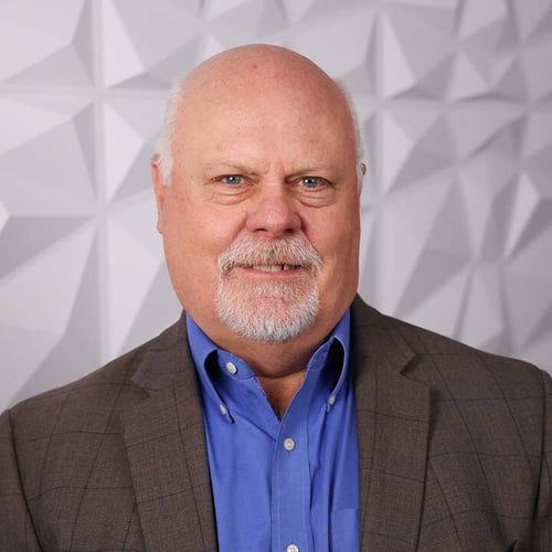 Portrait photo of UPAC member Scott Smith. He smiles at the camera in front of a white textured background. He is wearing a brown suit and blue shirt