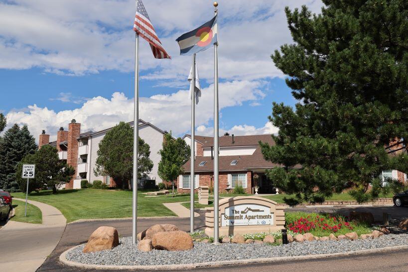 Apartment complex entrance pictured on a sunny day. A 2-way driveway is separated with a flower bed, gravel, & sign that reads "The Summit Apartments"