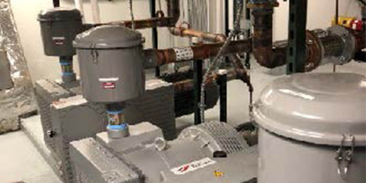 A photograph of copper pipes and grey pumps in a basement. The copper pipes run both horizontally and vertically out of frame. 