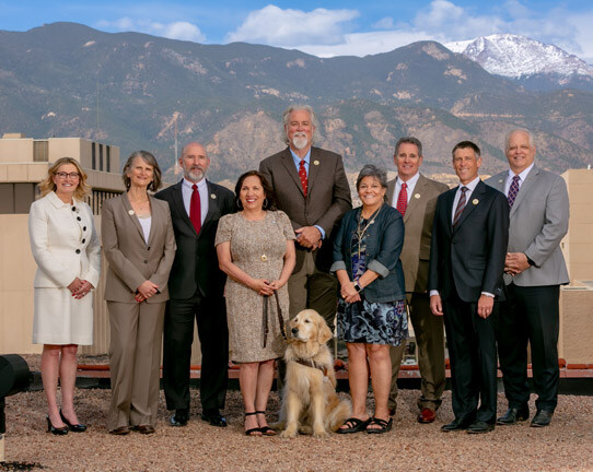 Group photo of the 9 current Colorado Springs City Council members wearing business attire on a downtown roof. Pikes Peak is visible in the distance.