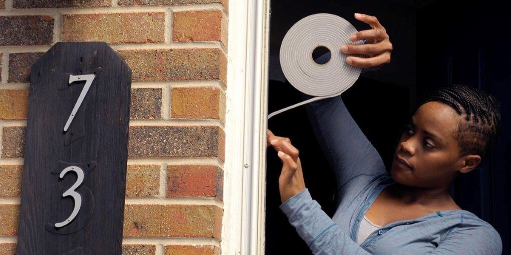 A woman installs weather stripping to her door frame for insulation. The house is brick and has a black address sign with the numbers "73" visible