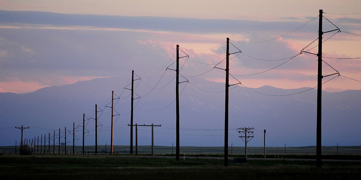 Photo of power lines in a large open field extending toward mountains in the distance. The sun is setting and the sky is blue and orange.