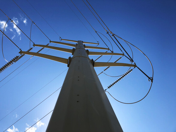 A photograph looking up from the ground at a large transmission pole and lines. The sky is blue.