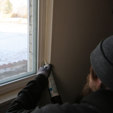 A man with a beard and wearing a black hoodie, gloves, and a beanie adds caulking to the seams of a windowsill. 