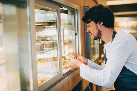 A person wearing an apron puts a cake in a refrigerator with glass windows in a restaurant setting.