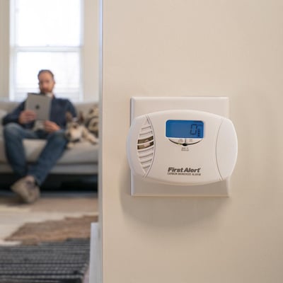 Carbon monoxide alarm mounted on a white wall. The meter reads as zero. Around the corner of the wall, a person reads a tablet on a couch. 