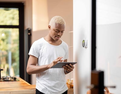 A person holding a tablet while standing next to a digital thermostat on a wall in a kitchen.