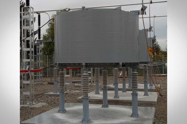 A series reactor in a substation. The reactor is a cylindrical shape. The scene takes place during the day time on a cloudy day.