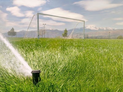 A sprinkler sprays water onto a soccer field. A goal is visible in the background with Pikes Peak visible in the distance.