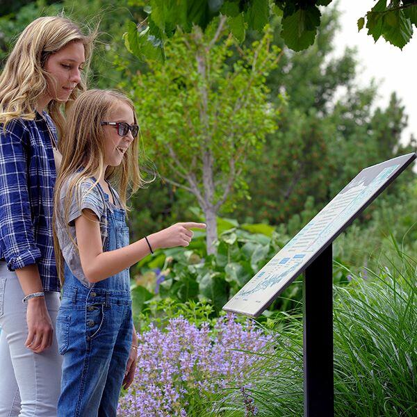 An adult and a child in an outdoor garden in front of an information plaque. The child is smiling and pointing at the plaque.