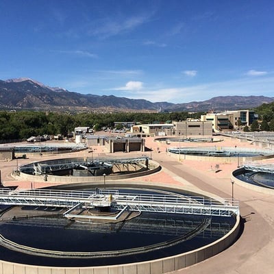 A wastewater recovery facility. Four circular basins are pictured on a sunny day with elevated walkways above them. There is dark blue water in them.